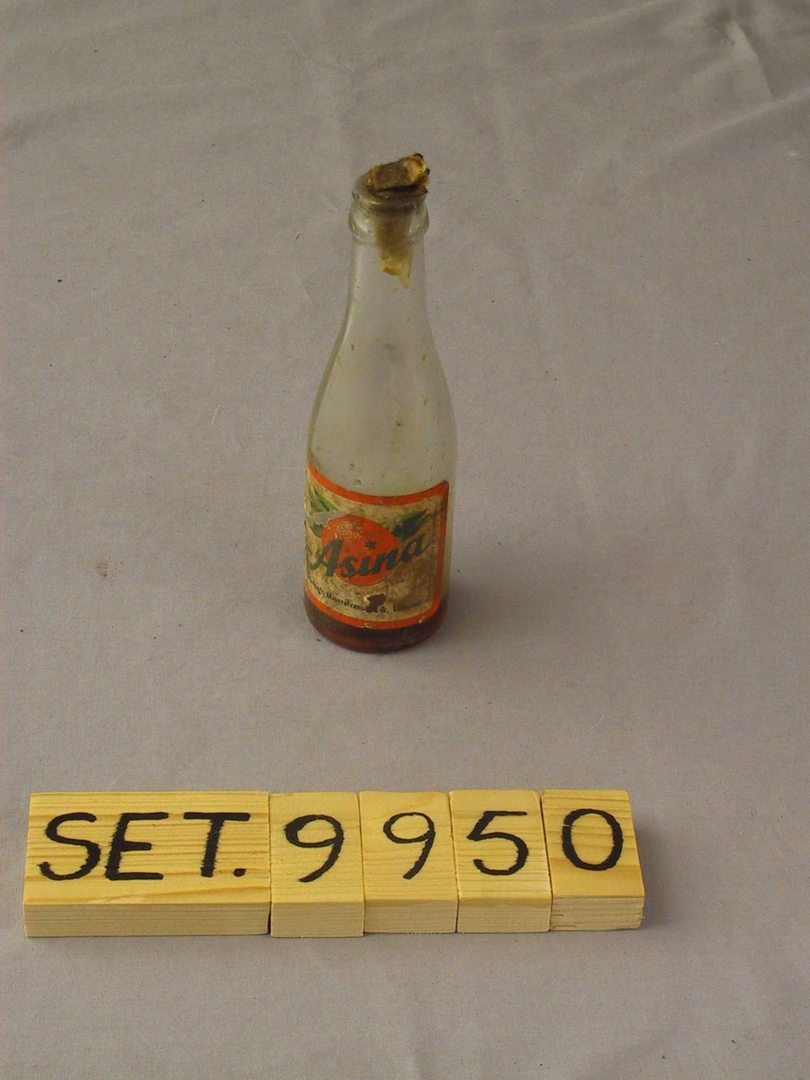 Glasflaske med jamn overgang mellom korpus og hals. Etikett. Inneheld liten skvett med olje,  "kork" av papir.