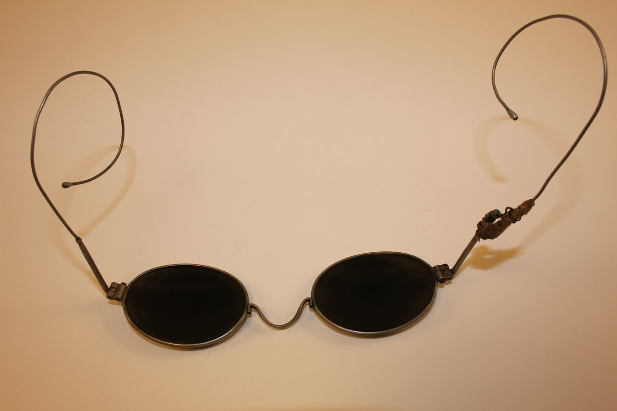 Beskyttelsesbriller/solbriller med ovale, sota glas. Lett bøyelege brillestenger. Venstre brillestong er knekt. Er reparert med hyssing og ståltråd. Brilleetui i papp med overtrekk av lêr. skinnklaff over opning.