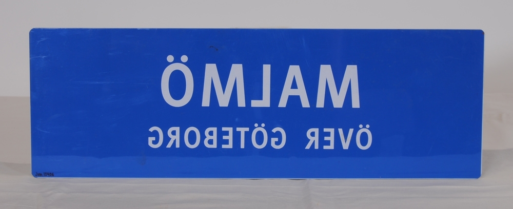 Destinationsskylt av ljusblå plast med texten "MALMÖ ÖVER GÖTEBORG" i vitt. Spegelvänd text på baksidan.