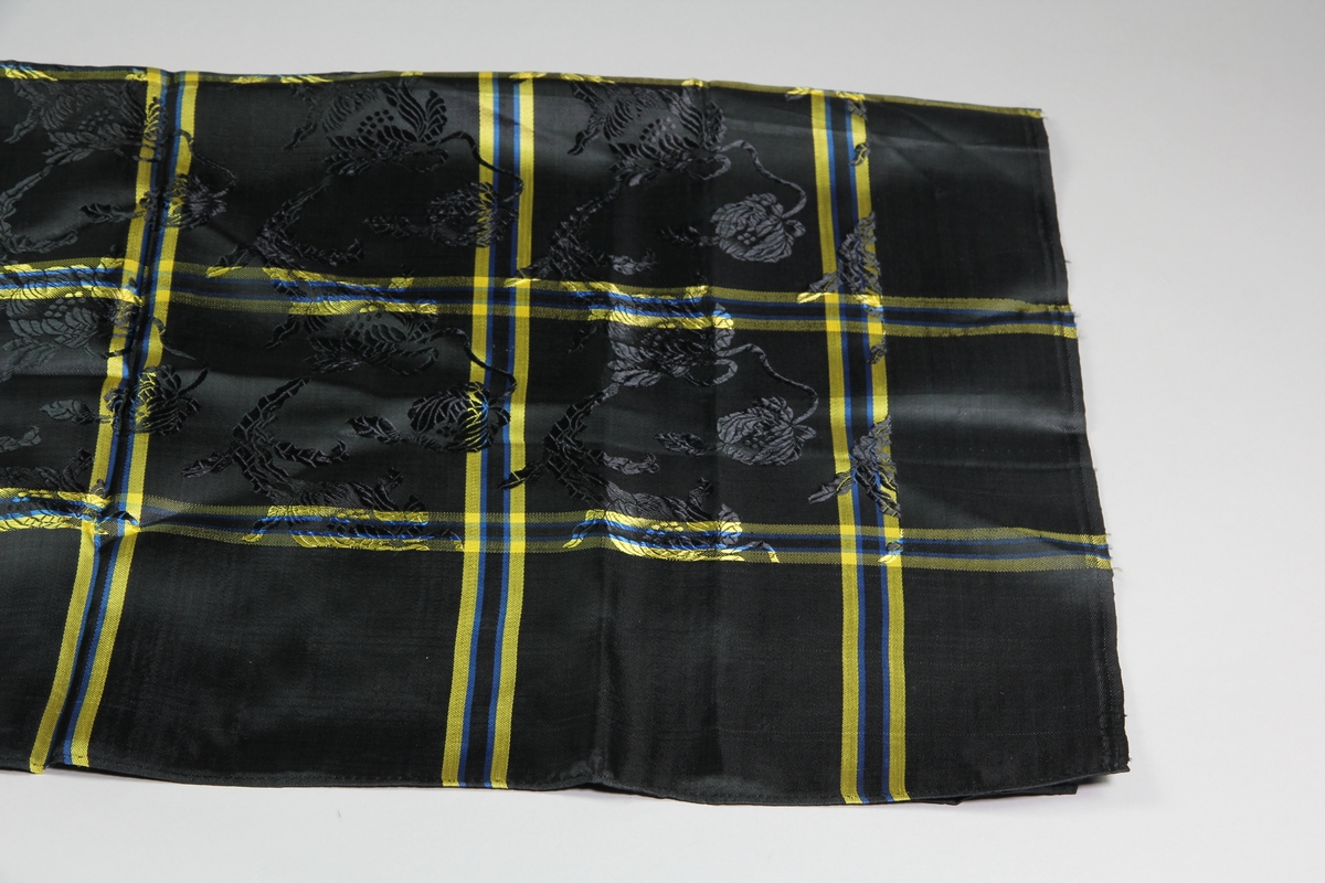 Kvadratisk svart sjal med gula och blå rutor i konstsiden. Den svarta botten har invävt blommönster. Smala handsydda fållar på två sidor, de andra två har stadkanter.