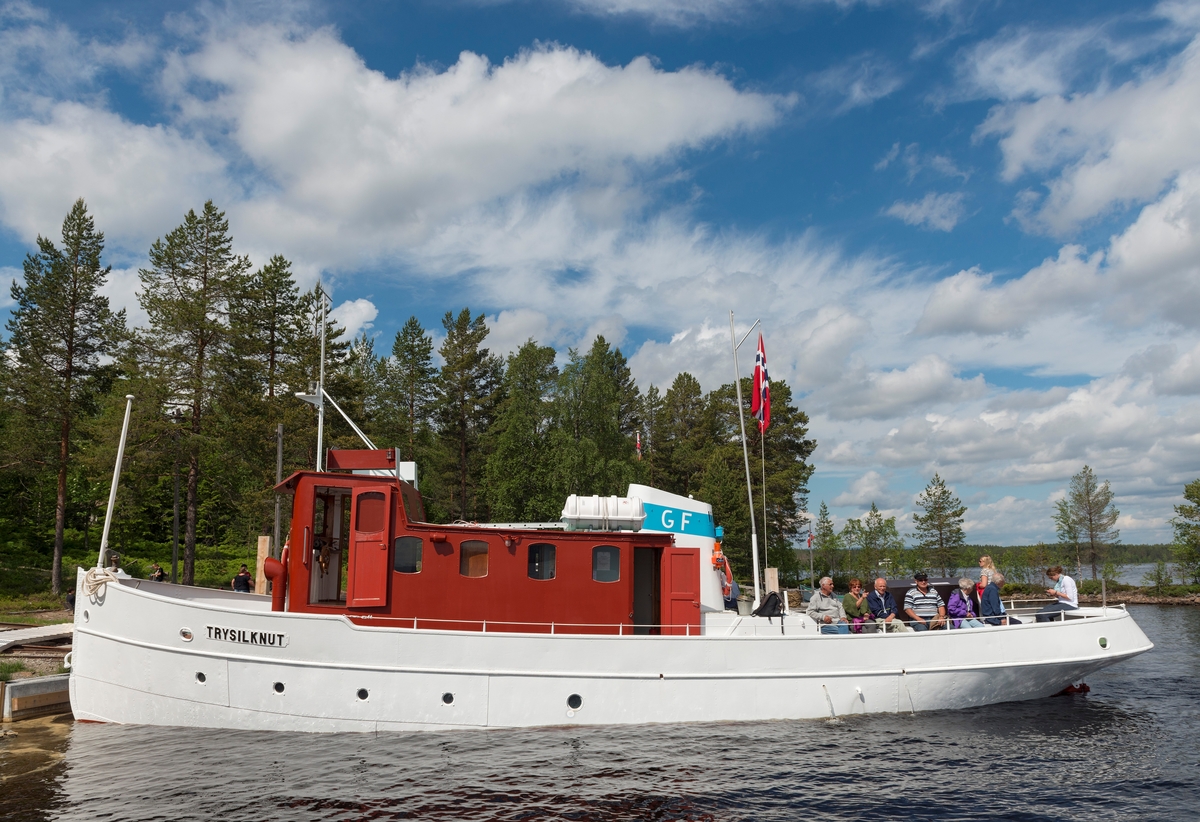 Slepebåten «Trysilknut» fotografert på den 47 kvadratkilometer store Osensjøen i grensetraktene mellom Åmot og Trysil i Hedmark, der Trysilknut gikk som tømmersleper fra 1914 til 1984.  Etter den tid har den 55 fot lange og 12 fot brede båten vært brukt som museumsbåt.  Den ble restaurert i perioden 2011-2014, og dette fotografiet er fra den første turen båten gjorde med passasjerer etter at restaureringa var avsluttet med maling av eksteriøret.  Skroget er kvitmalt og overbygningen (styrhus og bysse) er rødbrun.  Disse fargene har Trysilknut hatt siden båten ble ombygd fra damp- til dieseldrift i 1957-58, bortsett fra at vulsten som markerer hvor rekka møter den nedre delen av skroget lenge var grønnmalt og at Glomma Fellesfløtingsforenings initialer på skorsteinen skal ha hatt en gulere fargetone.  Fotografiet er tatt ved brygga på Sørlistøa fløtermuseum den 11. juni 2014, da museet markerte båtens 100-årsjubileum.  Mer om Trysilknut og dens historie under fanen «Andre opplysninger».