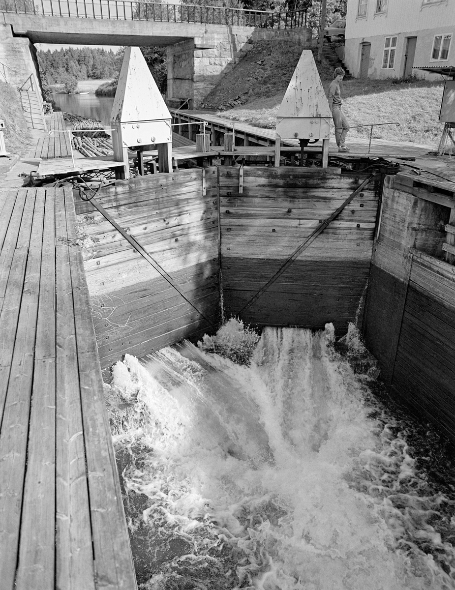 Den øvre sluseporten i Strømsfoss sluse i Aremark i Østfold.  Fotografiet er tatt høsten 1982, som var den siste fløtingssesongen i Haldenvassdraget. Fotografiet er tatt i motstrøms retning, med et nesten tomt slusekammer i forgrunnen.  Nederst på sluseportene ser vi hvordan kvitskummende vann fra overkanalen fosset inn i slusekammeret.  Dette ble i første omgang gjort for å «spyle ut» slusegodset – i dette tilfellet tømmer – fra slusekammeret og ned i underkanalen.  Når så nedre sluseport var stengt, ble de samme lukene brukt til å fylle opp igjen slusekammeret, slik at øvre port kunne åpnes.  Når så vannstanden i slusekammeret ble den samme som i kanalen ovenfor, kunne disse portene åpnes, slik at ei ny «slusevending» med tømmer kunne slippes inn.  Strømsfoss hadde bare dette ene slusekammeret.  Det utliknet en høydeforskjell på cirka to meter mellom den ovenforliggende Strømselva og den nedenforliggende kanalen mot Aremarksjøen.  Ettersom trafikken her var stor, prioriterte Haldenvassdragets Kanalsekskap å mekanisere betjeninga av dette sluseanlegget.  Så vel sluseportene som lukene fikk hydrauliske åpne- og lukkemekanismer, som ble aktivert via brytere i et skap ved høyre bildekant.  Da dette fotografiet ble tatt var det Anne Johansen som betjente dette bryterpanelet.

En liten historikk om tømmerfløting og kanaliseringsarbeid i Haldenvassdraget finnes under fanen «Opplysninger».