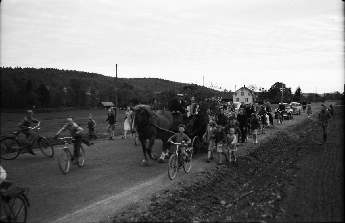Kortege på landsvägen med lövad vagn och dragspelare. Kusken heter Ingemar Sjögren och enligt uppgift tilldrar sig händelsen "vid flytt av Sjuttas vid Strängbetong". Många cyklar och i bakgrunden syns även några bilar.