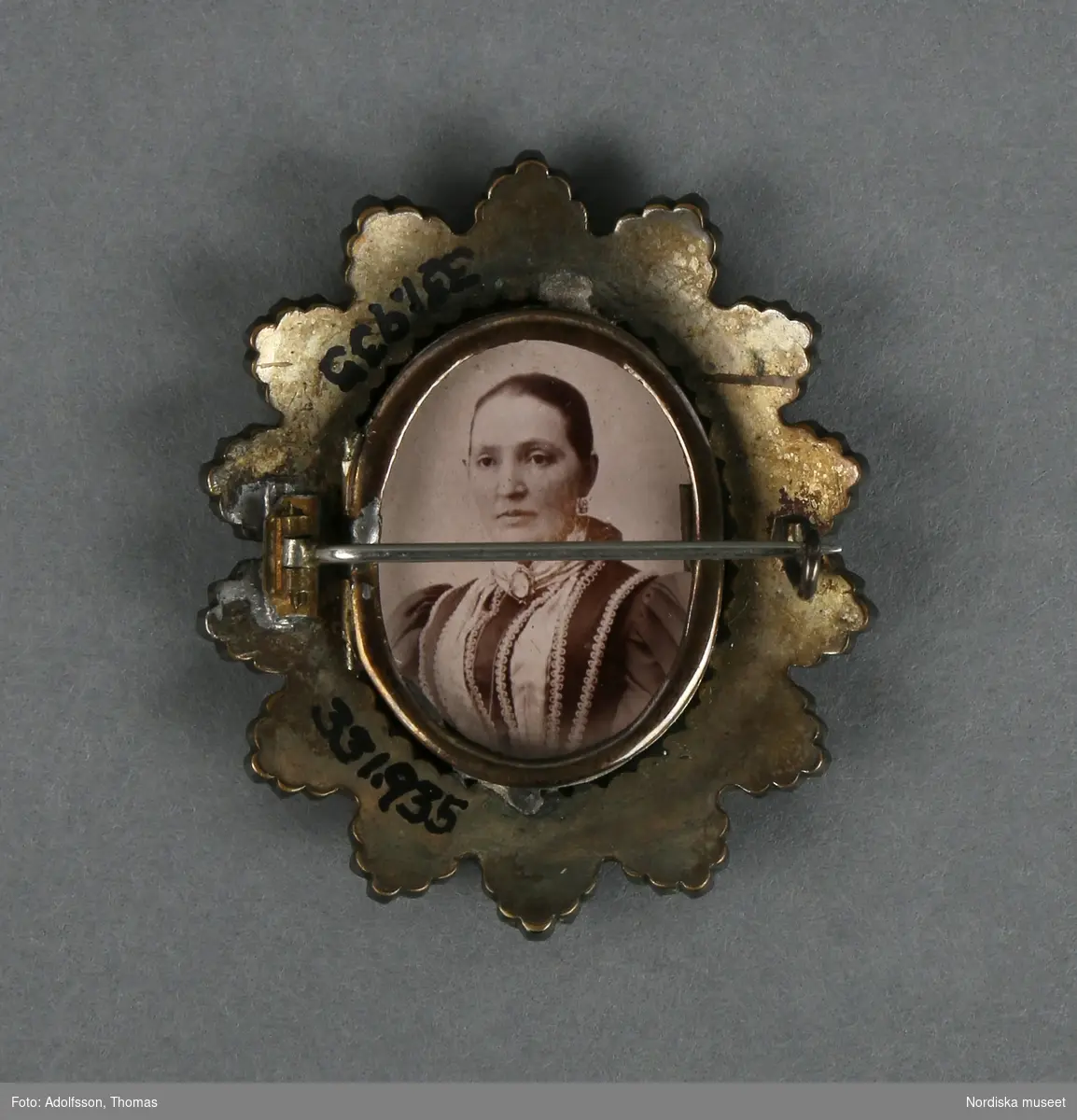 Brosch av gulmetall och granater (eller rött glas) med fotografier. Framsidan med uddformig krans klädd med granater (?) och innanför en smal ram av gulmetall med fotografi bakom glas.   Baksidan med nål och hake, samt smal oval ram (likadan som på framsidan) med foto. Fotografierna som det ena föreställer en man och det andra en kvinna, (trol. tagna på 1880--1890-talet) är lösa och kan byta plats med varandra, beroende på vilket foto man vill ha mot fram, respektive baksida. 
Ramen på baksidan är öppningsbar. 
Anm. några av granaterna saknas. Glaset till fotot på baksidan saknas
/Leif Wallin 2014-06-24