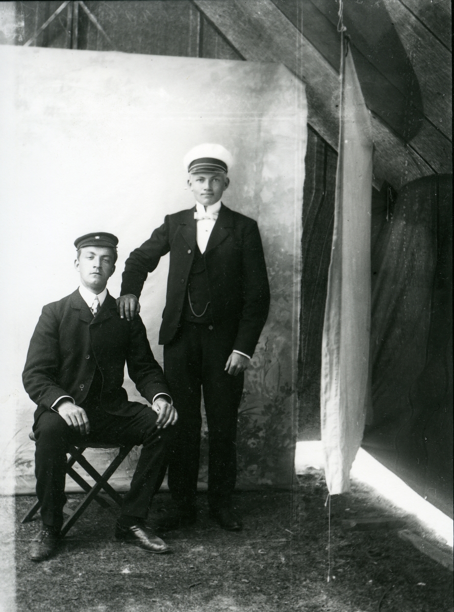 To unge menn er avbilda foran eit lerret. Begge er kledd i dress med kvit skjorte og hatt på hovudet. Den eine guten sit på ein stol medan han andre står ved sida av med den eine handa på skuldra til den andre.