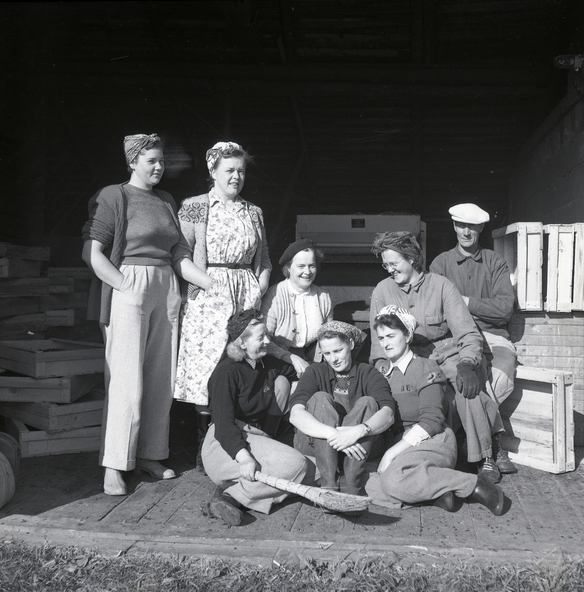 En grupp kvinnor med sjalar runt håret och en sopkvast sitter framför en hög med trälådor. Där sitter även en man med keps.
