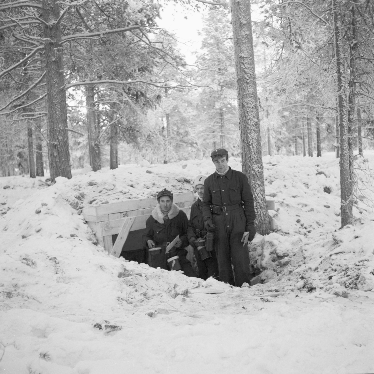 Tre militärer vid nedgången till ett markvärn efter utspisning. I händerna håller de bröd och matkärl. Vid Svenska frivilligkåren i Finland, F 19.
