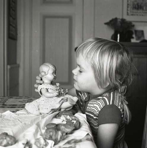 En flicka sitter vid ett bord och leker med en docka, 1972.