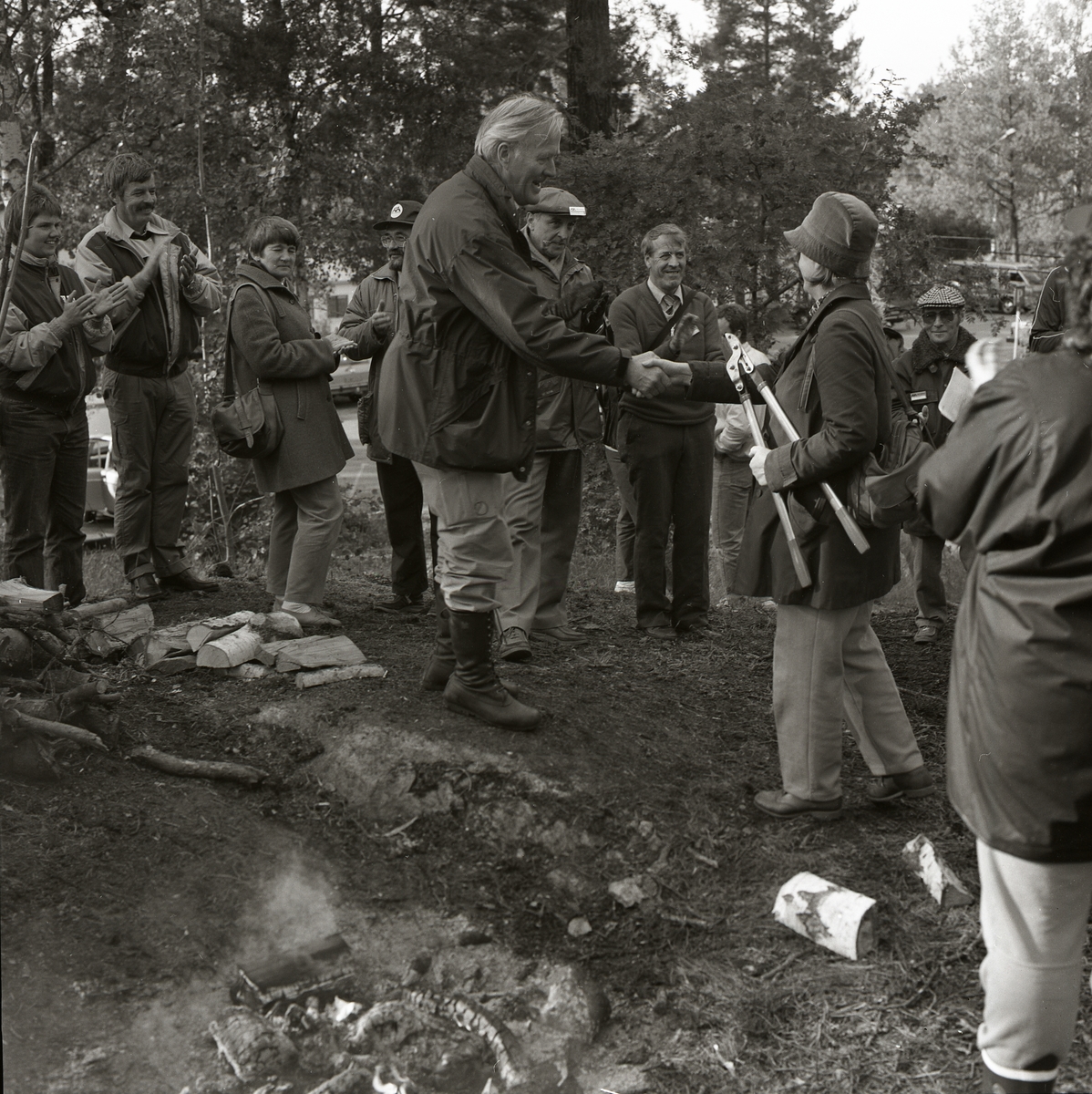 Mellanskog, Sånga-Säby, 28-29 september 1985. En man och en kvinna tar varandra i hand. Kvinnan håller i en grensax. Runt dem står en grupp människor och applåderar. På marken ligger ved och det brinner en brasa.