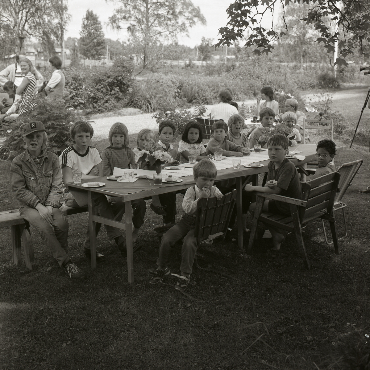 En grupp barn sitter runt ett trädgårdsbord under ett midsommarfirande på gården Sunnanåker den 25 juni 1983.