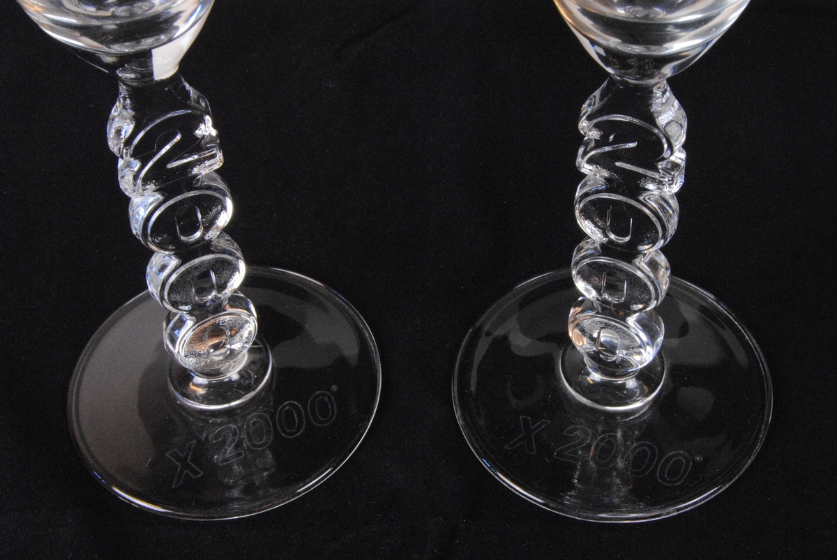 Två champagneglas av pressglas i flûtemodell med tulpanformad kupa. Stjälken är utgörs av texten "2000" med siffrorna staplade på varandra. Foten är cirkelformad och har texten "X 2000" ingraverad i kursiv.

Jvm23606:1 och :2 är identiska.