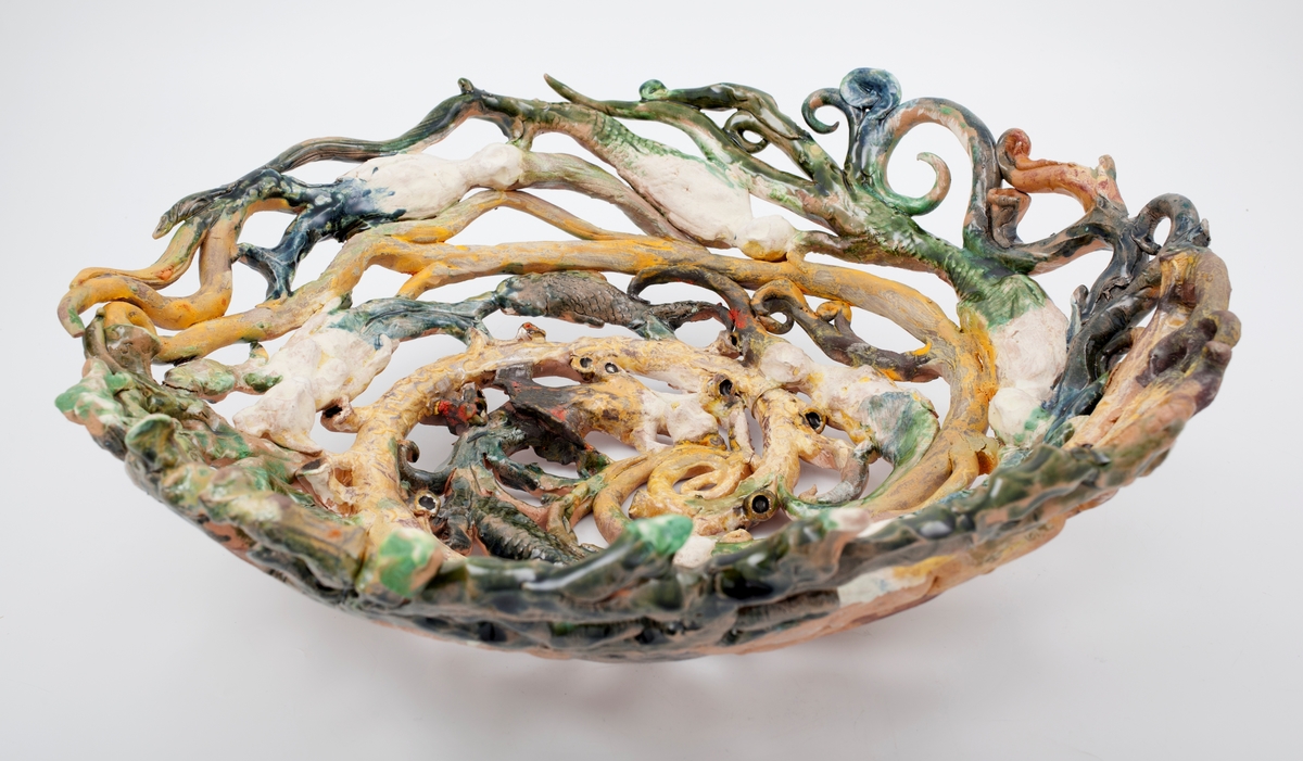 Stort, polykromt gjennombrutt keramikkfat. Fatet er bygget opp av spiraler og dekorert med havmenn og havfruer.
