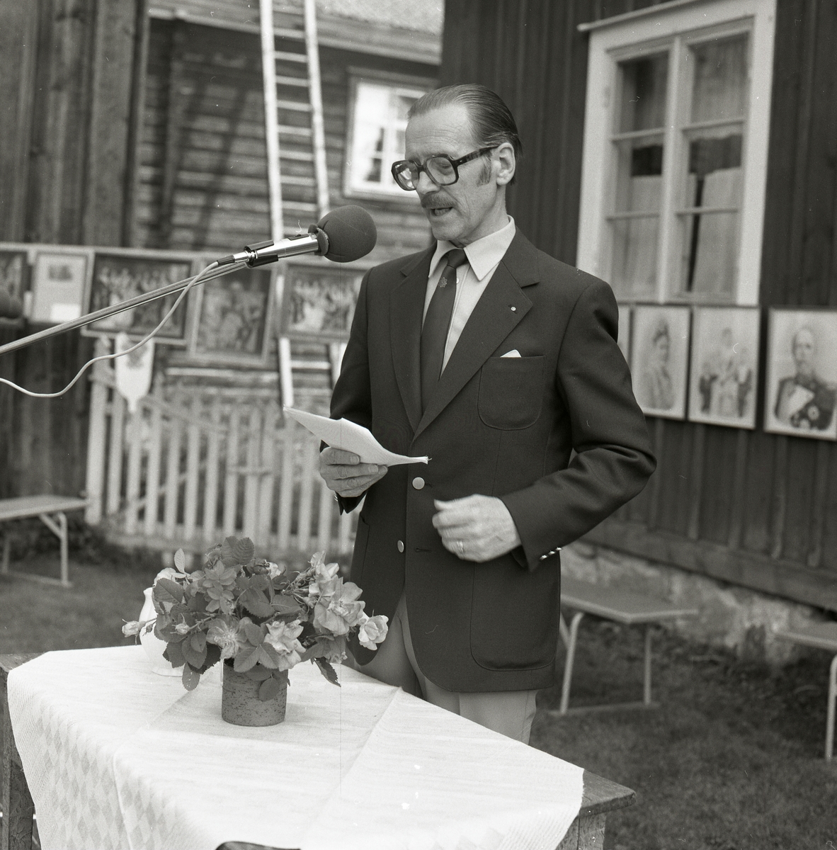 En man iförd kostym, slips och glasögon står vid en mikrofon och läser ett tal. Framför honom står ett bord med duk och blomkruka. Bakom honom hänger tavlor på husväggen, Hembygdsbyn 1981.