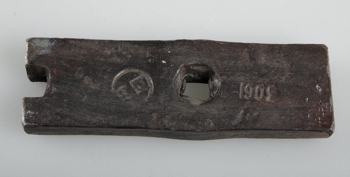 Avhugget stycke av järnband, byggnadsjärn, med ett och ett halvt fyrkantigt hål, stämplat med en rund stämpel EB och 1902..