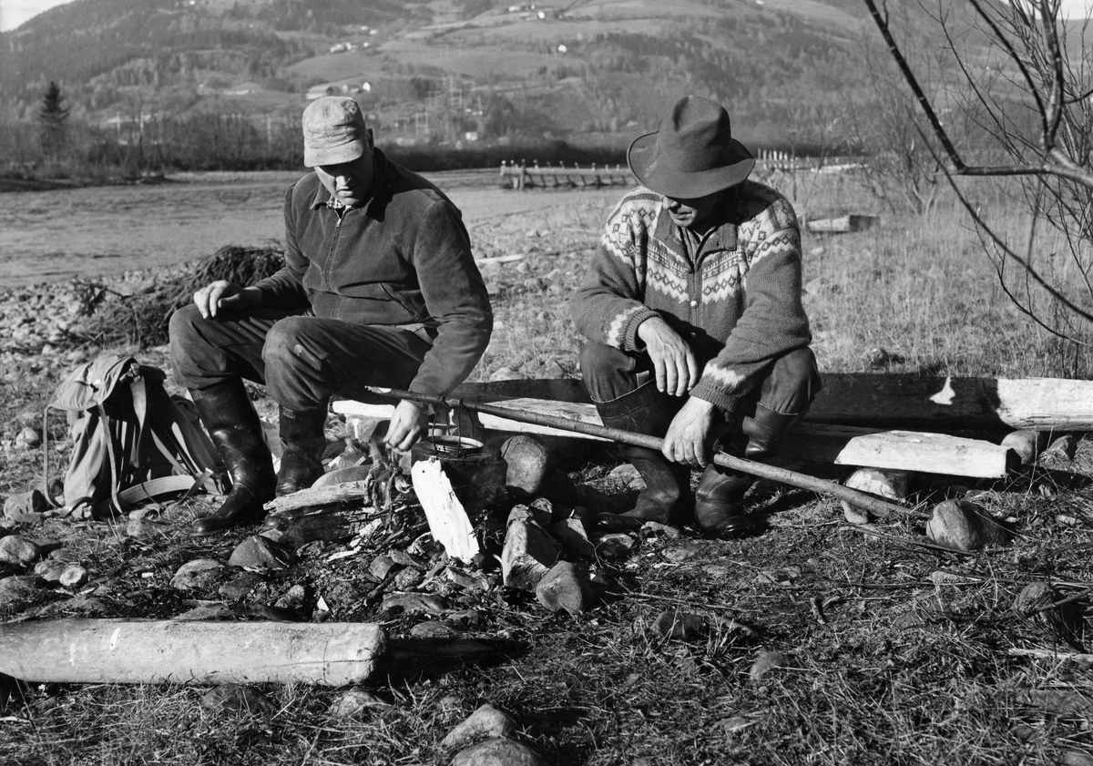 To fiskere, fotografert ved kaffevarmen ved elvebredden på Øyra i Fåberg høsten 1962.  En av dem holdt en sotete kaffekjel over varmen på ei stang som han støttet mot en stein som sto på høykant.  Karene var arbeidskledde.  Mannen til venstre satt ved sida av ryggsekken sin.  I bakgrunnen skimter vi et lendings- eller utgjerdstre som er montert som strømbryter i elveløpet for å lette notfisket.
