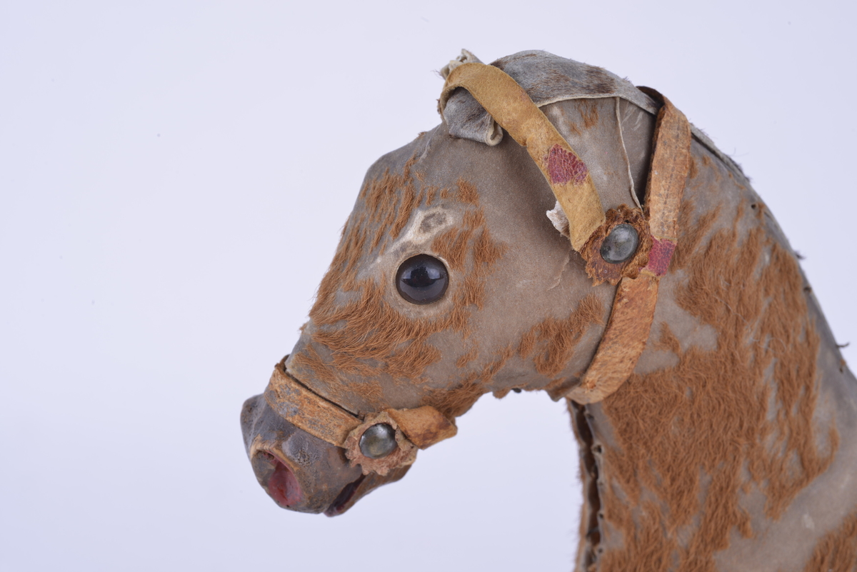 Modell av hest montert på en rektangulær treplate med fire hjul av tre. Hesten er utformet av skinn og fyllt med spon. På utsiden av skinnet er det limt på imitert hår/tekstil. Sal og bissel av lær.