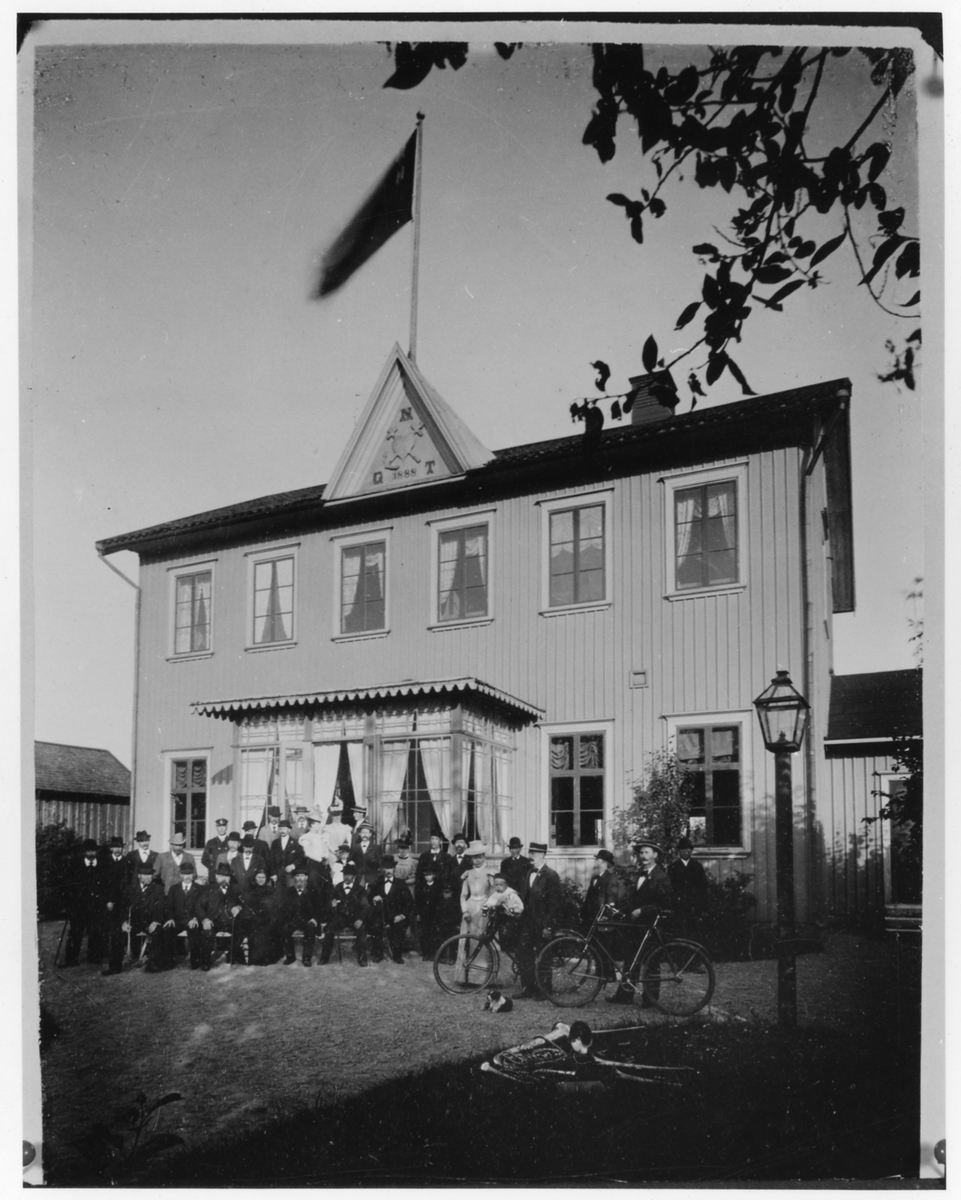 Gruppbild av 34, sittande och stående, kvinnor, män och pojkar framför ett hus. Huset har ett tympanon med texten N, G, T 1888 samt en symbol av ett hjärta korsat med kors och ett ankare. Till höger på bilden ses två cyklar, musikinstrument på gräset och ett småbarn. Stående vid cykel t.h. Fridolf Thorssin. 

NTO:s lokal i kv Hammaren vid Göteborgsvägen.