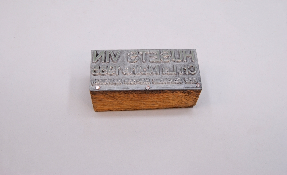 Rektangulär kliché av silverfärgad metall, monterad på en träkloss. På klichén finns texten: "HUSETS VIN Ch. La Mothe 1955 Röd Bordeux Hel-och Halvbutelj", i releif.