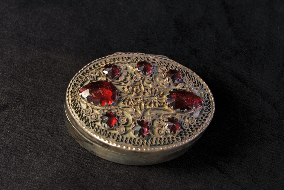 Oval dosa av silver. Locket är prytt med filigranarbete och infattade, slipade glasstenar (8 st.) i rött. Allmogestil. Stämplad invändigt i botten.