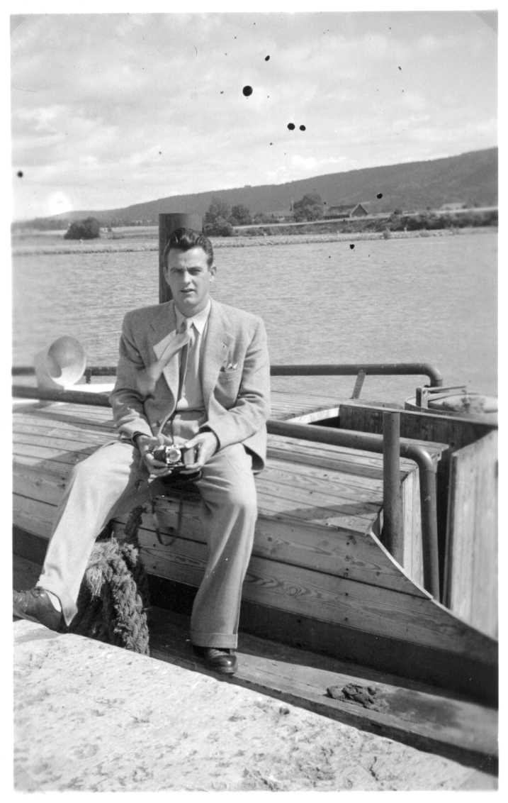 En ung man i kostym och slips med kamera i handen sitter på en båt i Gränna hamn.