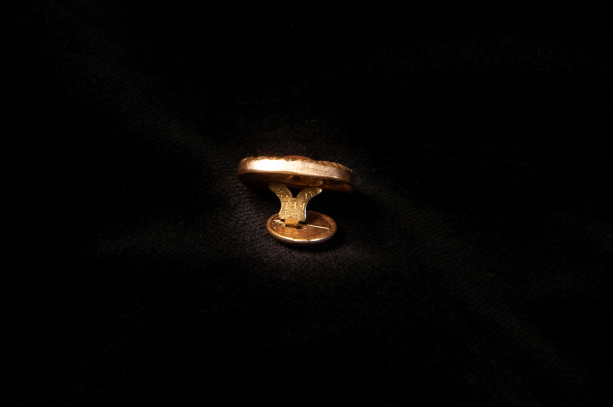 Fyrkantig stolpknapp av förgylld metall (gulddoublé). Präglad dekor i form av zickzack-bårder m.m, och en infattad rund pärlemoknapp 
(troligen imitation) med ett hål i mitten.