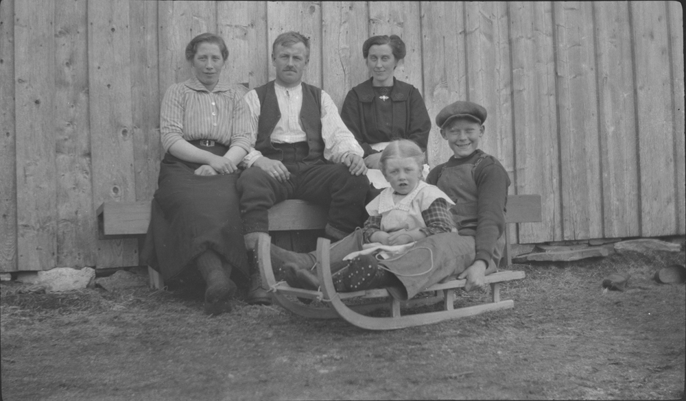 Tre voksne sitter på en benk ved husvegg, jente og gutt sitter på kjelke foran.
Fra v. Ingrid, Lars og Mathilde Øyen. På kjelken: Nelly Øyen (g. Fjellseth) og Einar Karstensen.