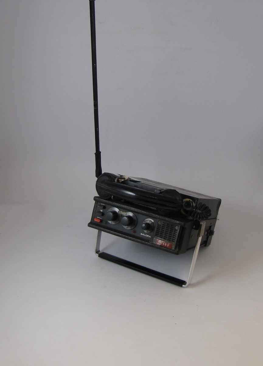 En svart rektangulær gjenstand med et håndtak, en antenne og et telefonrør. Det er tre knapper og tre vridbare knotter på en konsoll på toppen av gjenstanden, på siden av røret.