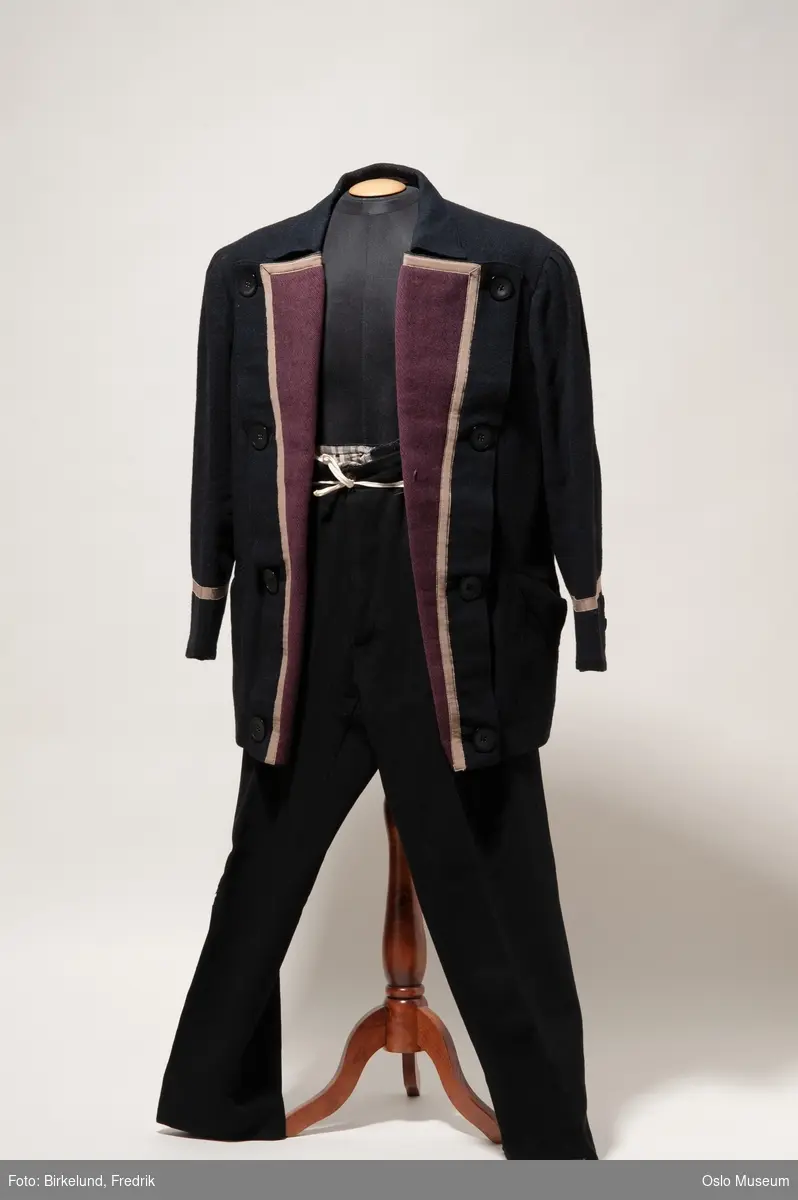 A: uniformsjakke, sort ull med brune slag og lysebrunt silkebånd. Store sorte knapper på slagene.
B: sorte ullbukser