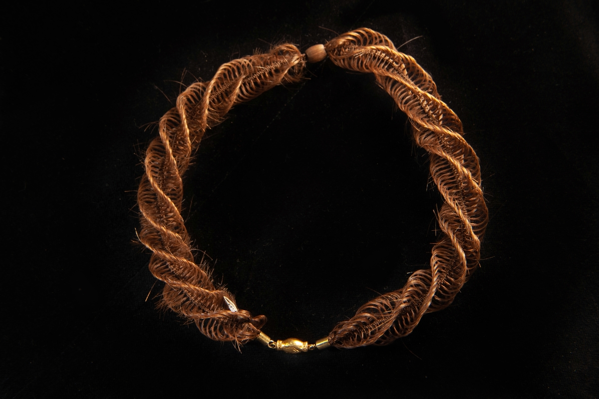 Ett vridet halsband av brunt hårarbete, flätat i bågar och spiraler med en spunnen kula mitt fram. Guldlås med pressad dekor i form av två händer som möts.