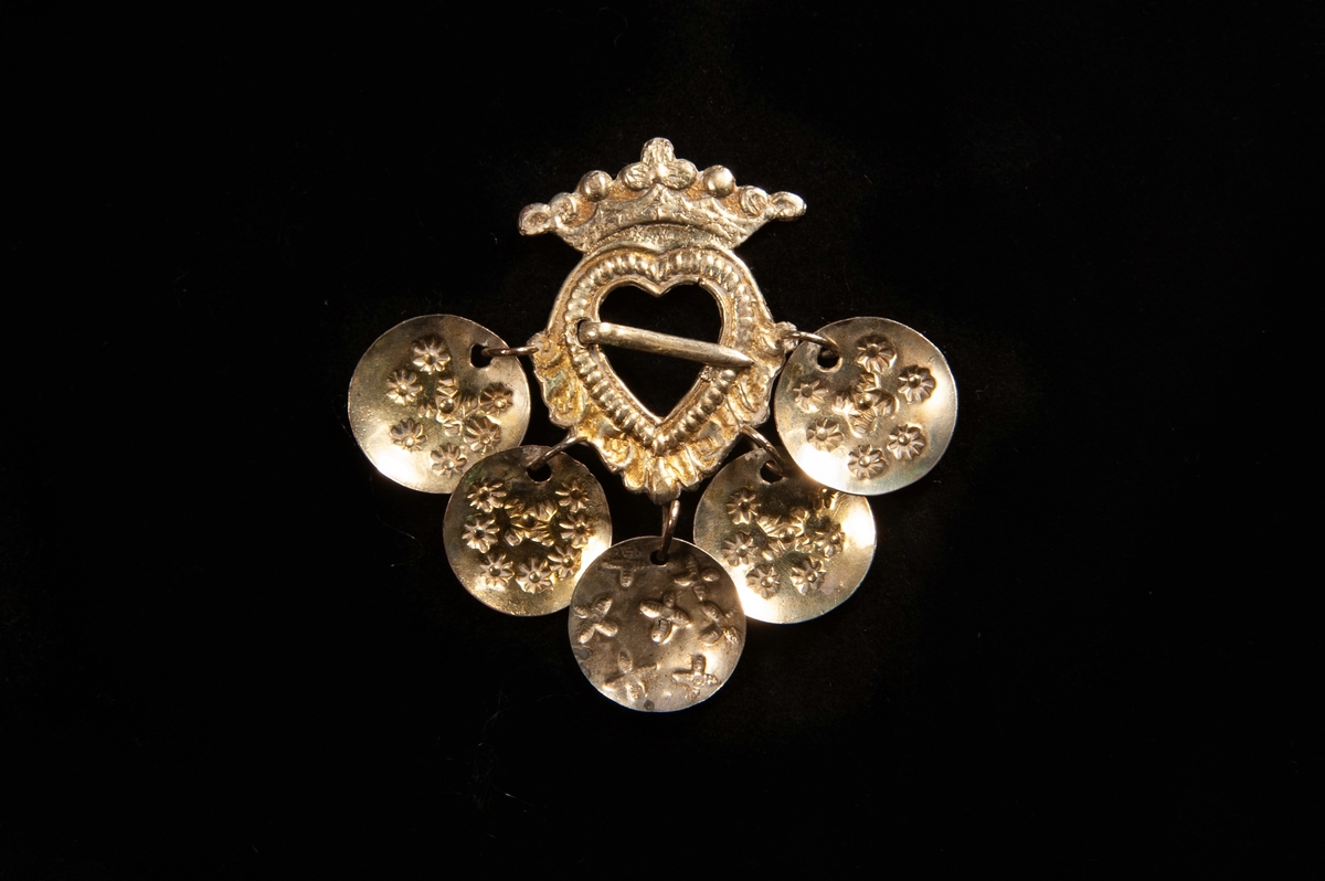 Hjärtformad bröstsölja av graverat och förgyllt silver, med nål. Krona överst och fem mönsterstämplade löv av silver hänger ner från spännet. Saknar stämplar.