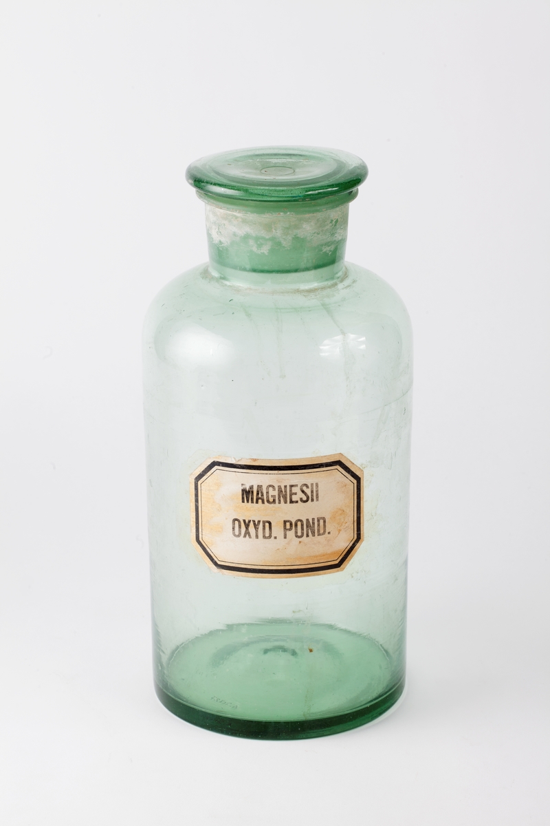 Standglass med slepet glasskork til oppbevaring av pulver i laboratoriet. Gul etikett med sort ramme.