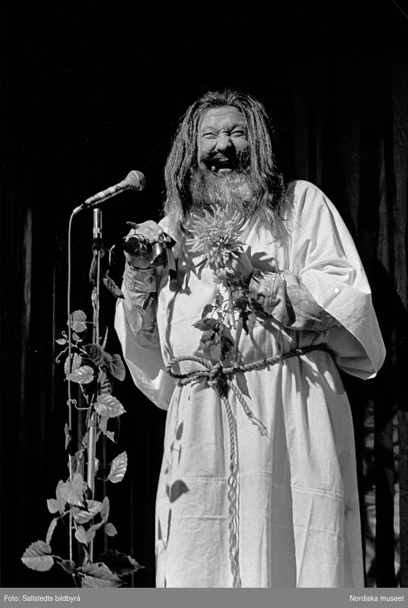 Skådespelaren Carl-Gustaf Lindstedt framträder på Berns i en peruk med långt hår, långt skägg, vit särk med rep om midjan hållande blommor. Utstyrseln liknar klichébilden av en 60-tals guru.