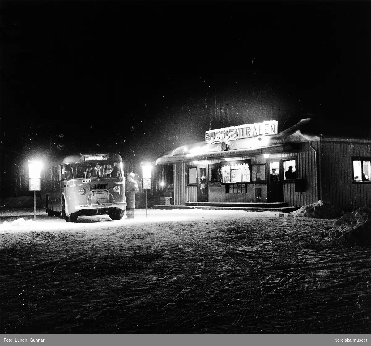 Nattbild, en buss står parkerad vid Busscentralen, Rättvikstrakten år 1951. Bussen är en Scania årsmodell 1946. Marken är täckt av snö.