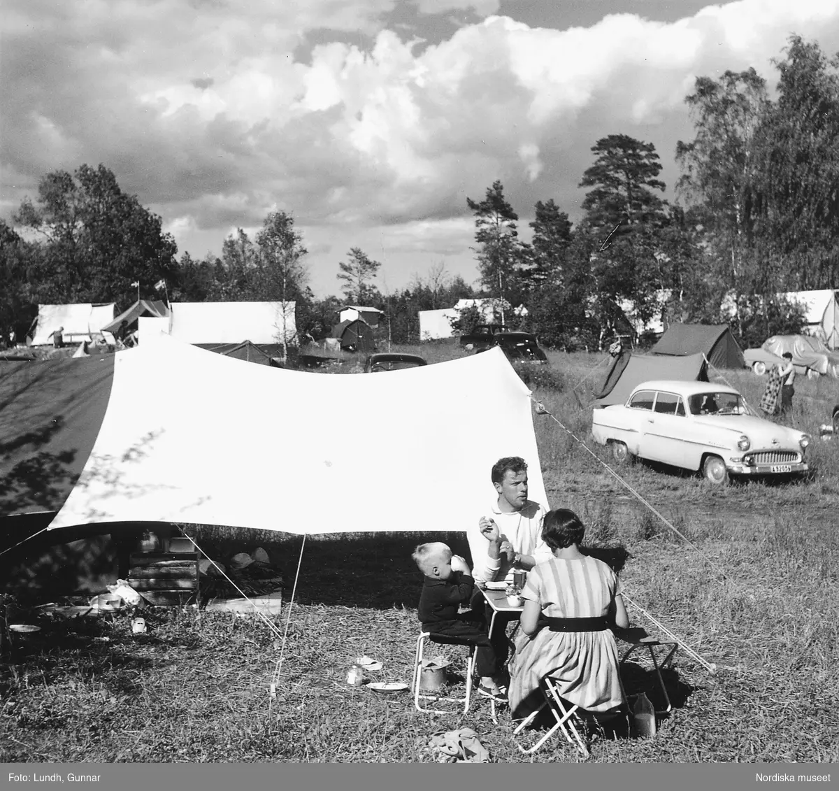En campingplats med tält och bilar på en äng i skogen år 1956. En man, kvinna och barn sitter vid ett campingbord och äter. Bilen till höger är en Opel Rekord.