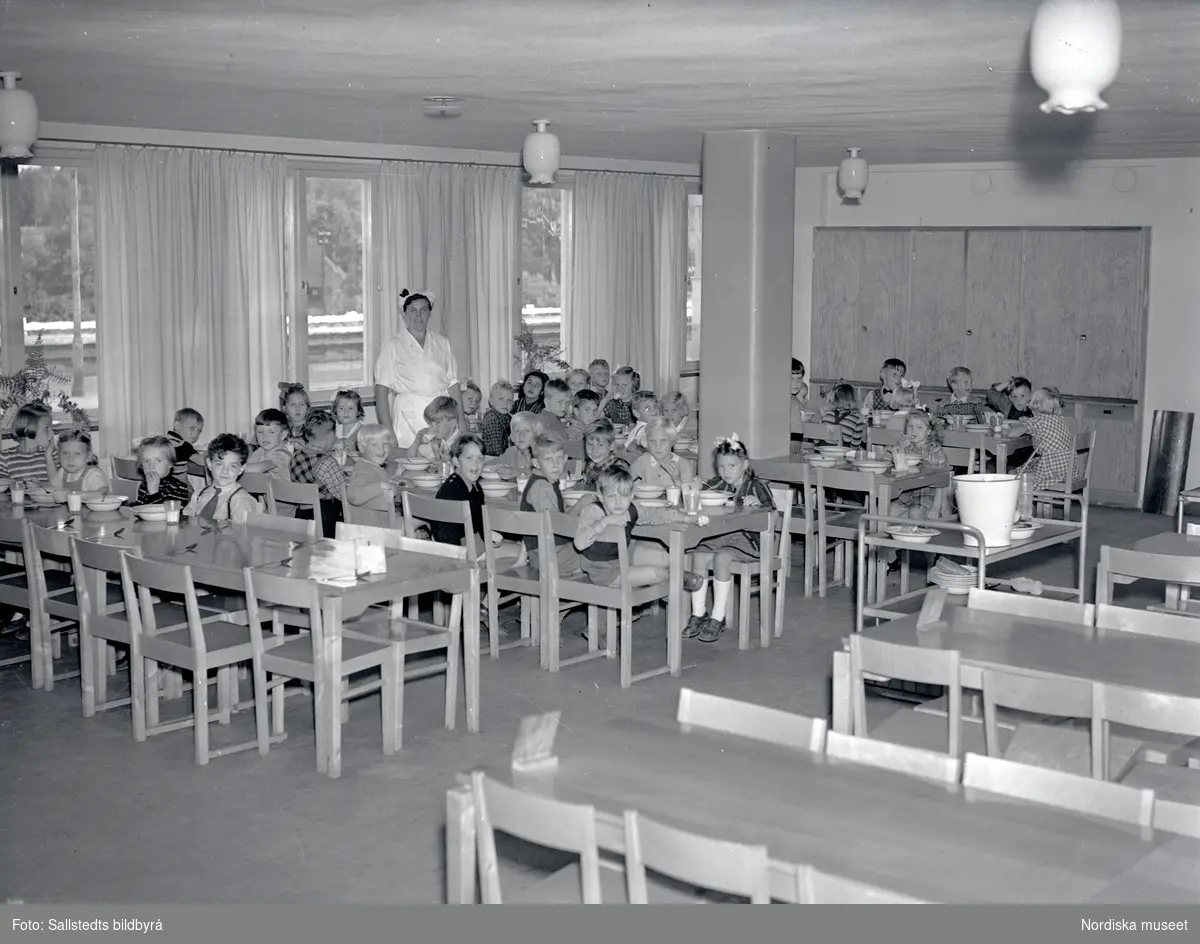 Barn som tittar på fotografen sitter i en barnbespisning på Solna folkskola år 1946. En kvinna i vita arbetskläder står till vänster i bild. Bilden är tagen för att användas i valreklam.