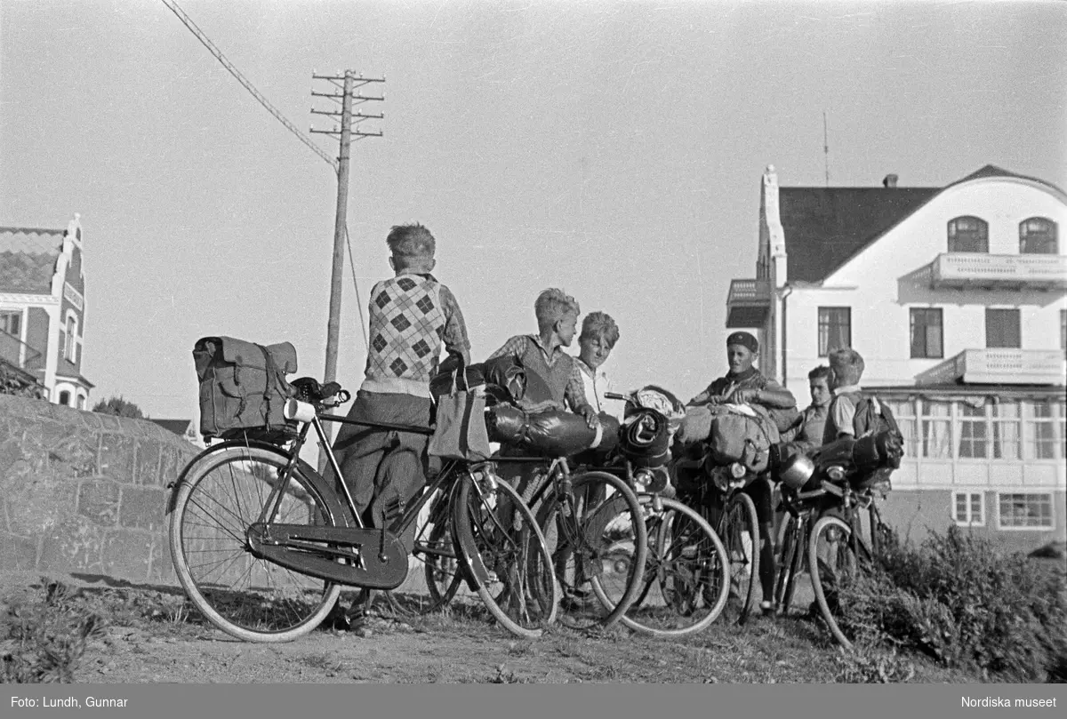 En grupp pojkar med cyklar står vid vägen utanför Grand Hotell i  Mölle ( i bakgrunden t.h.), Cyklarna är lastade med ryggsäckar och sovsäckar,