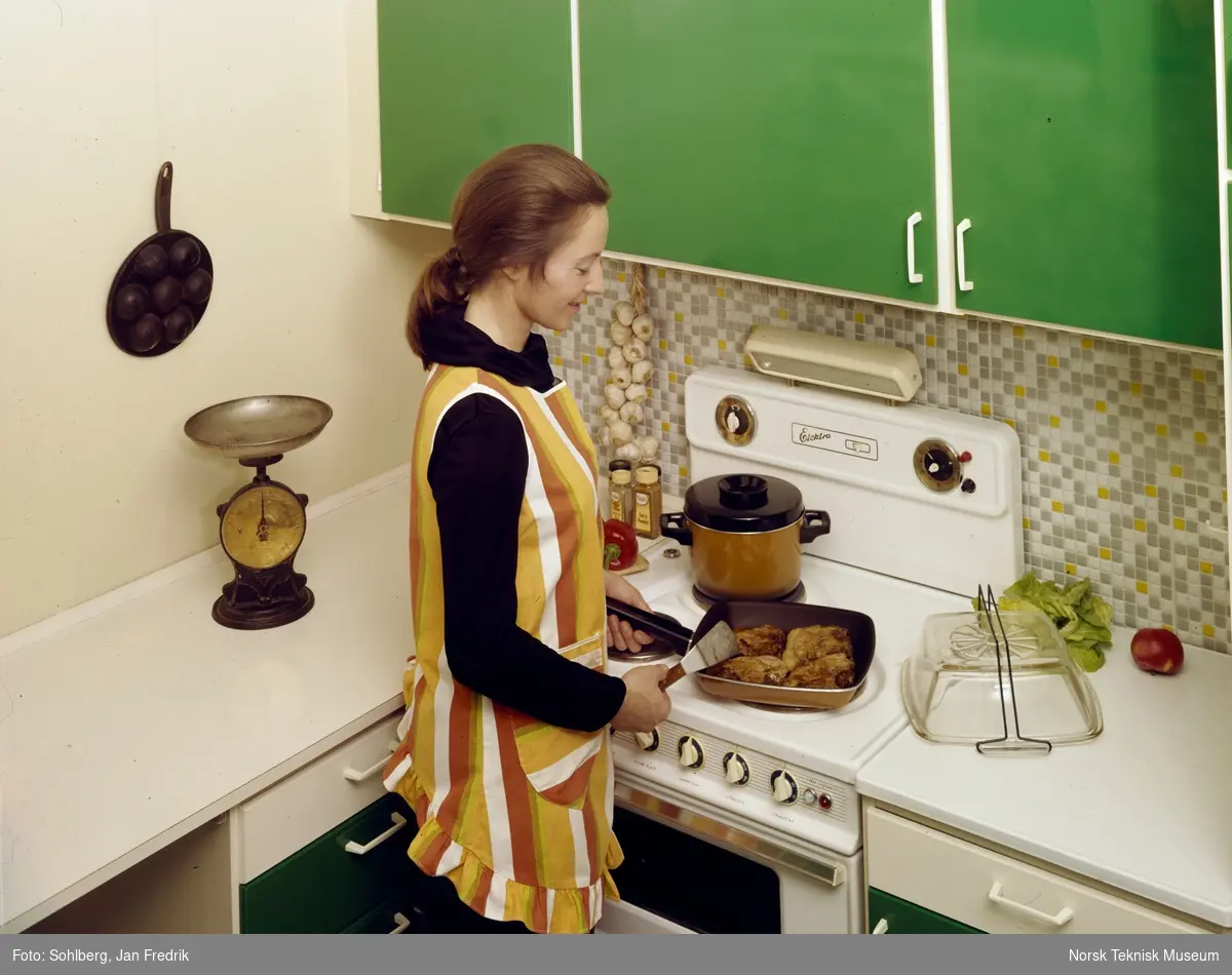 Reklamefoto for komfyr Elektra. Med moderne kjøkkenutstyr er livet som husmor ganske enkelt. En smilende kvinnelig modell lager mat ved komfyren.