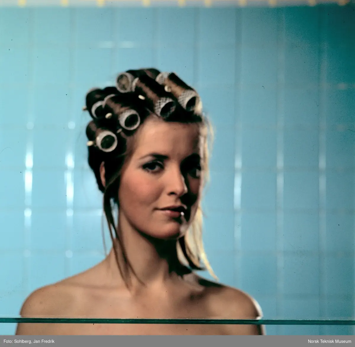 En kvinnelig modell viser fram tidens hårmote. Hun har håret oppsatt med krøllspenner. Turkise fliser i bakgrunnen. Bar overkropp.