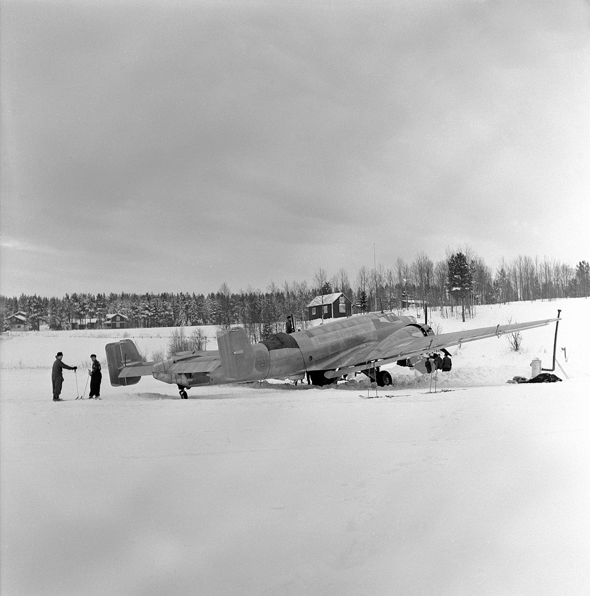 Flygplan B 3 nödlandad på Storsjöns is efter flygning i hårt väder i februari 1946. Skidåkare vid flygplanet. Vy snett bakifrån.