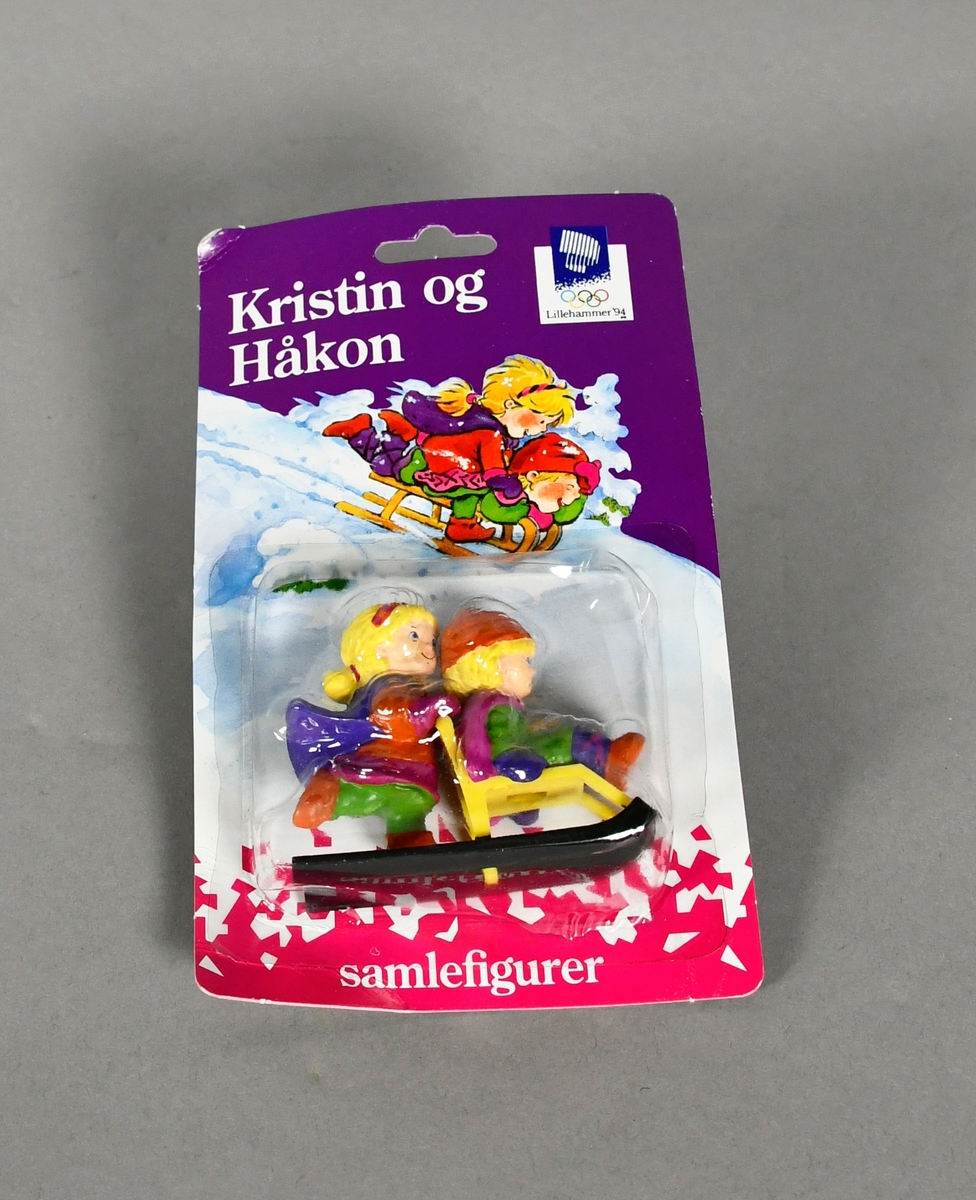 Samlefigur som fremstiller maskotene Kristin og Håkon, der Håkon sitter på en spark og Kristin sparker. Figuren er pakket i original emballasje.