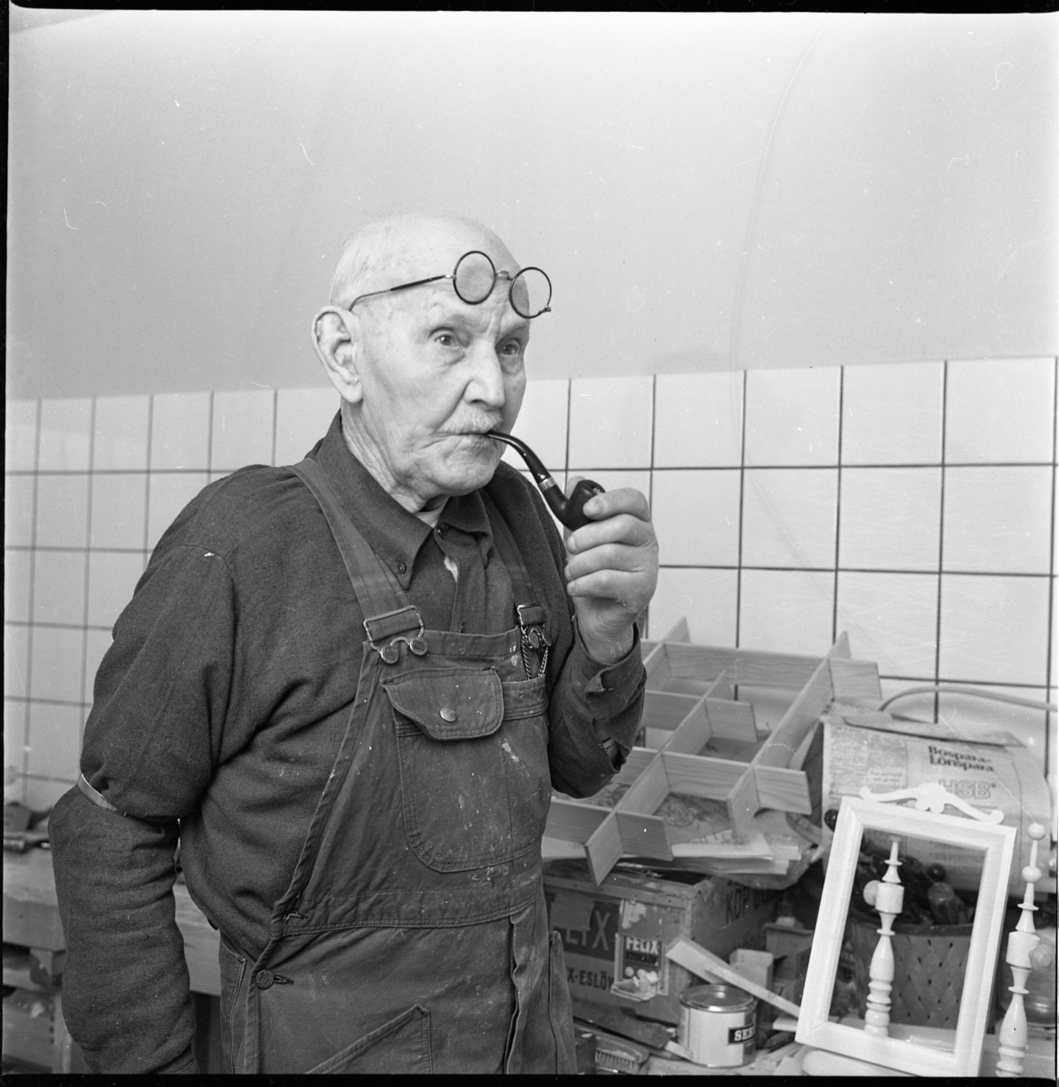 Korgmakaren Emil Lund, 91 år, står i snickarbyxor med runda glasögon uppdragna i pannan och pipa i munnen.