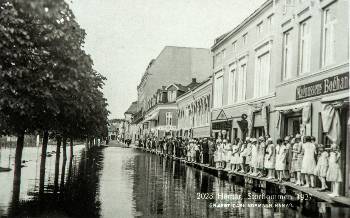 Postkort, Hamar by, flom i Strandgata, Mjøsflommen 1927, folk går på gangbroer utenfor Magnussens bokhandel, 