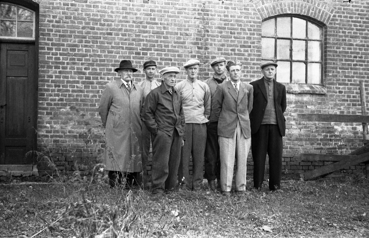 Sju uidentifiserte menn avbildet foran en teglsteinsvegg. Bildet, som er fra høsten 1956, kan muligens være fra potetmelfabrikken/brenneriet på Lena.