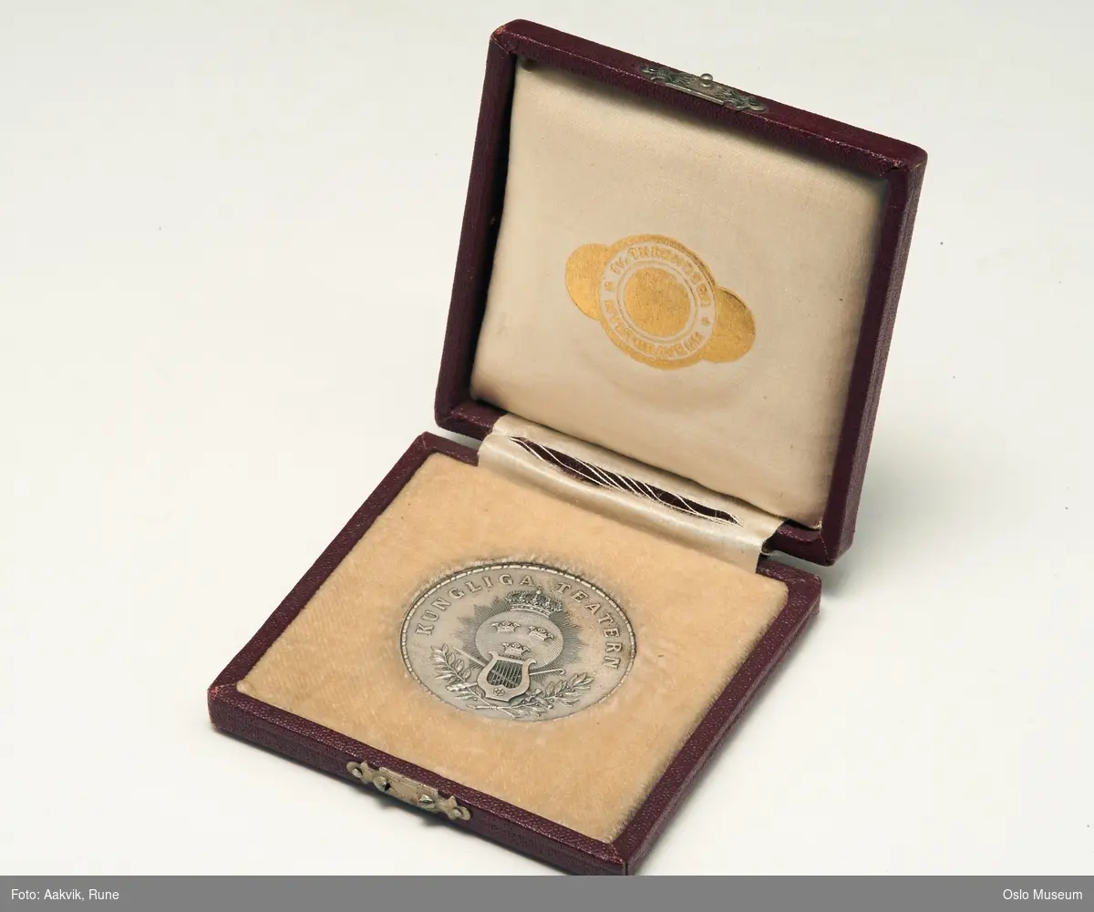 Sirkelrund medaljong i etui, sølv med innskruft og moriv med "tre kronor", lyre, laubærgrener (?), krone, etui har ødelagt lås