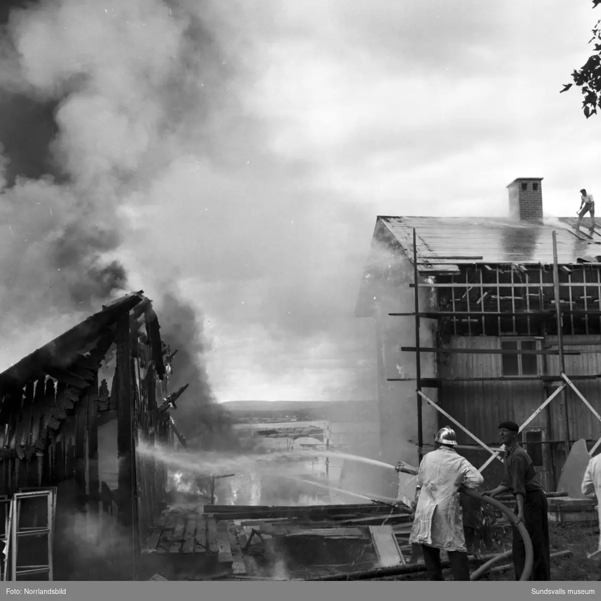 Släckningsarbete vid en eldsvåda i en uthuslänga i Skönvik. De boende bär ut bohag ur det närliggande bostadshuset.