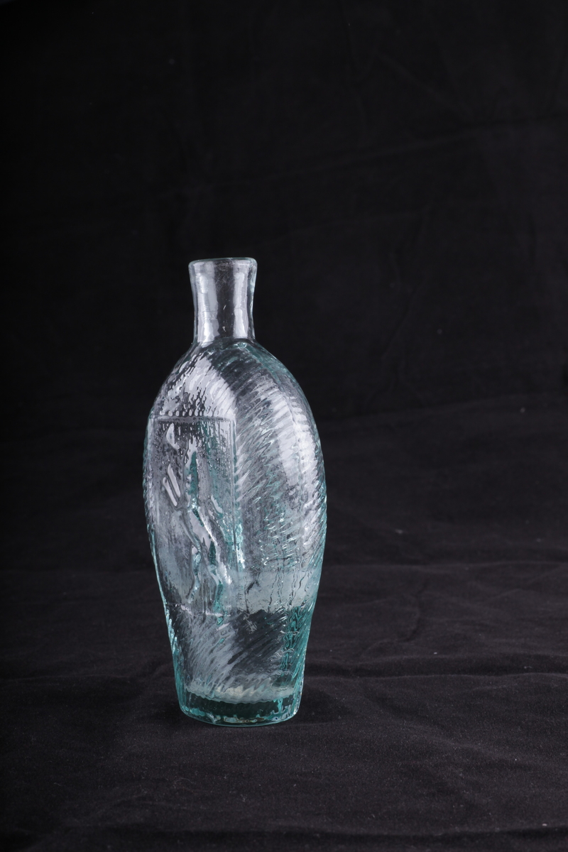 Flaske i grønnlig glass, blåst i form. Rommer en halv pegel. Motivet på den ene siden viser den norske løve, på den annen en drueklase.