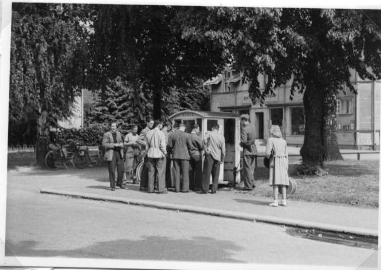 Ett antal män stående vid en glasskiosk på okänd ort. Till höger en militär uniform och en kvinna. I bakgrunden ett trähus med skylt om blanda annat Bok- och pappershandel.
