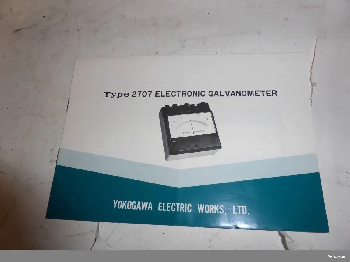 En galvanometer i en svart läderväska. I en plastficka som sitter fastsatt i väskans bärrem finns det en manual på engelska, en lapp med instruktioner på svenska, och en extra manual för en mätspänningskälla. Galvanometern är tillverkad av Yokogawa Electric Works, Ltd. i Japan.