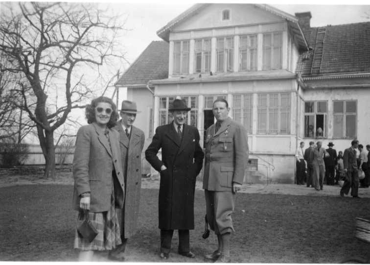 En grupp flyktingar, män och kvinnor i trädgården till Hamnvägen 3. I förgrunden en kvinna med solglasögon samt tre män varav en i uniform.