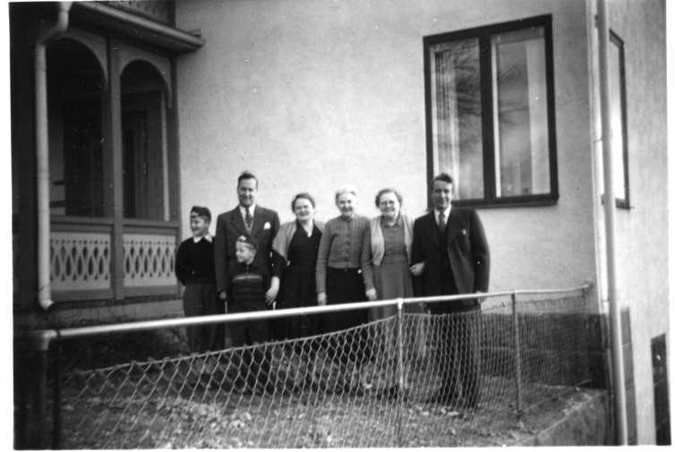 Tre kvinnor, två män och två yngre pojkar stående i ett led tätt intill varandra intill ett hus.